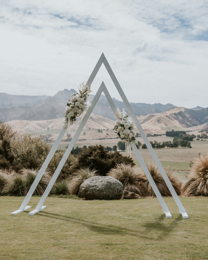 Triangular wedding arch with floral decorations in a scenic outdoor setting with mountains and greenery.
