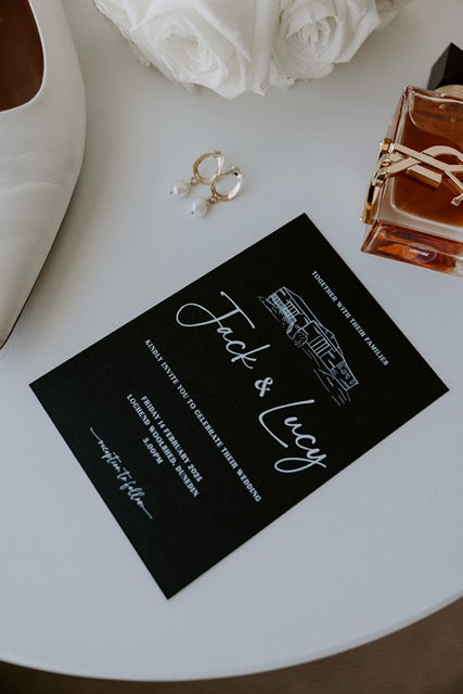 Black wedding invitation card with white ink and custom illustrations on a light surface.