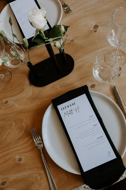 Table setting with white plates, black menus, and floral arrangements on a wooden table.
