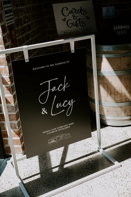 Wedding welcome sign for Jack and Lucy against a brick wall with wooden barrels.