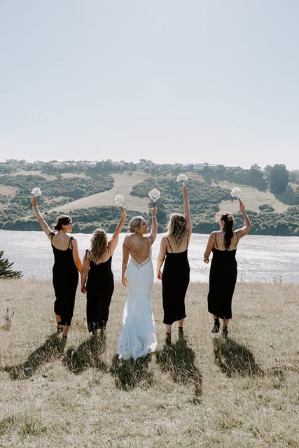 Group of women in black dresses with one in a white dress, standing on a grassy field with a scenic background.