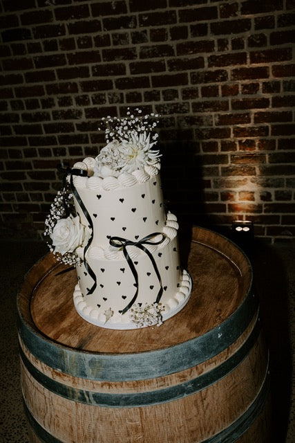 modern wedding cake with white flowers and black ribbons on a wooden barrel against a brick wall.