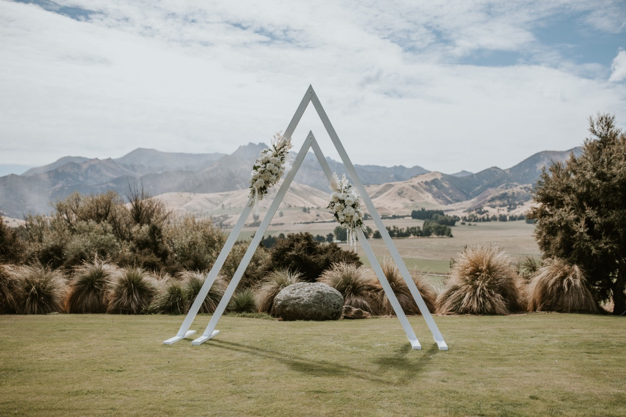 Triangular wedding arch with floral decorations in a scenic outdoor setting with mountains and greenery.
