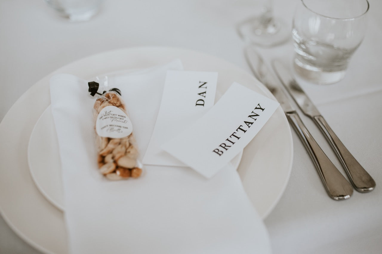 Table setting with name cards for 'Dan' and 'Brittany', a small bag, and cutlery on a white tablecloth.