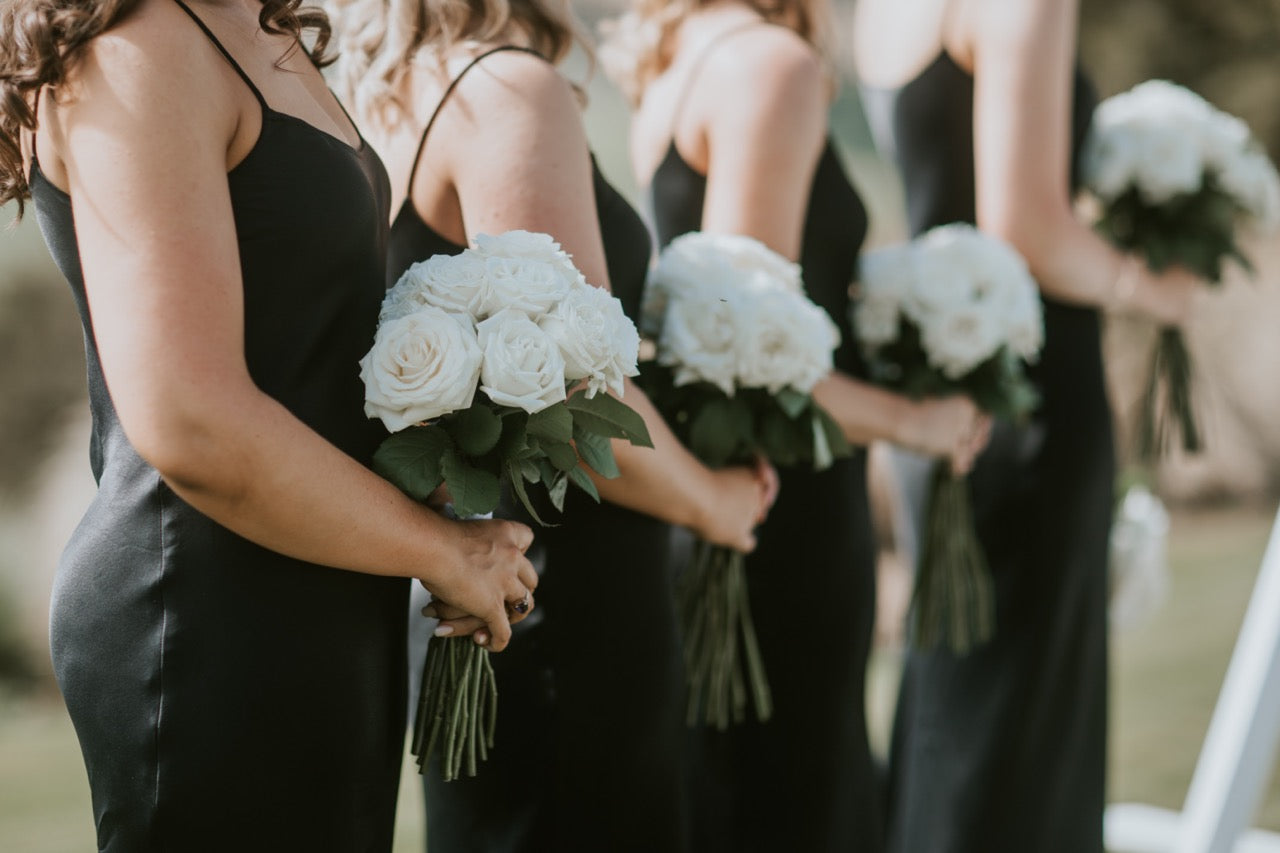 Bridesmaids in black dresses holding white rose bouquets at a wedding.