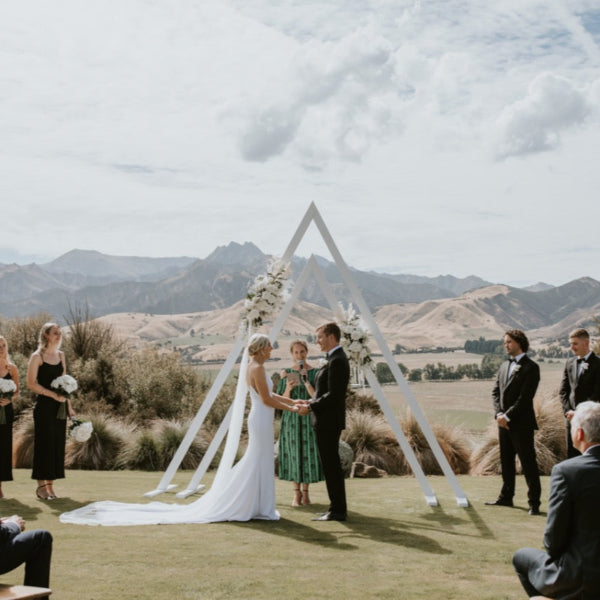 Wedding ceremony with a double white triangle arch on a grassy area with mountains in the background at the Lookout Lodge Wanaka