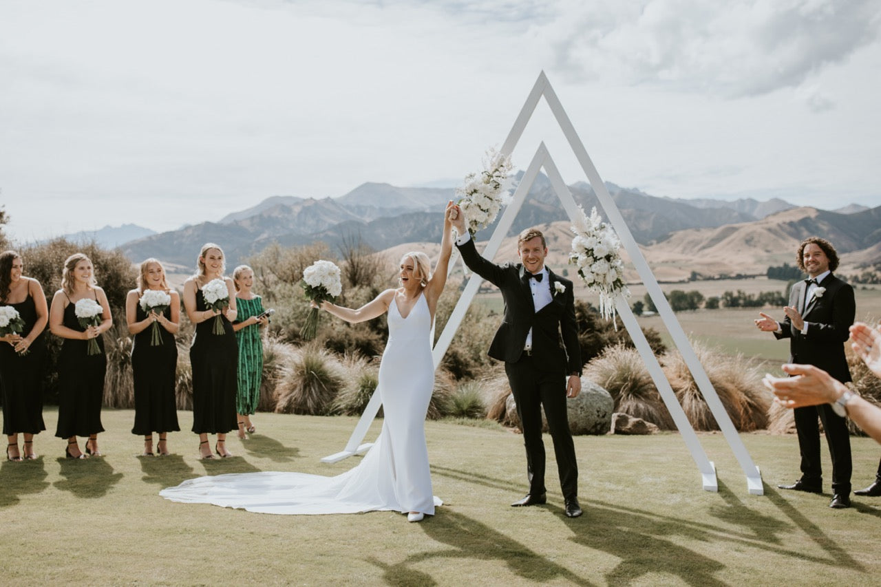 Wedding ceremony with a double white triangle arch on a grassy area with mountains in the background at the Lookout Lodge Wanaka