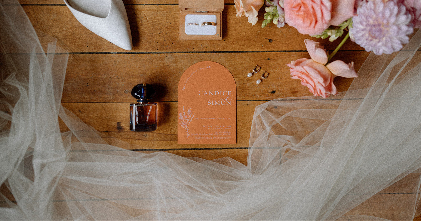 Orange wedding invitation with white ink  and floral arrangement on a wooden surface.