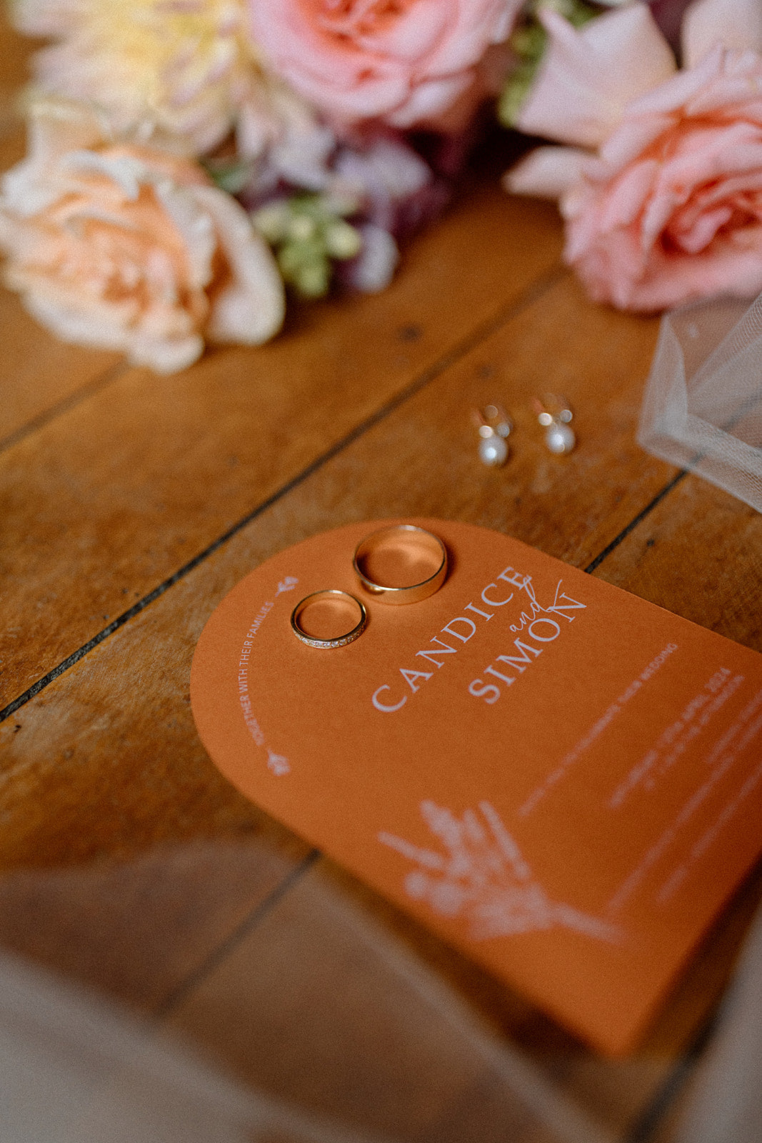 Orange wedding invitation with white ink  and floral arrangement on a wooden surface.