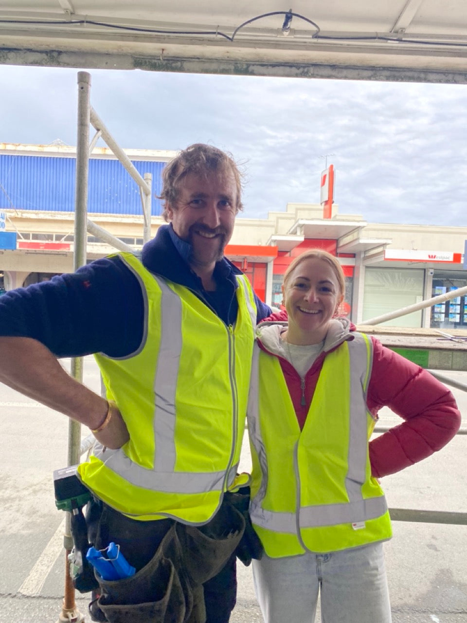 Two people smiling and laughing wearing high-visibility vests doing a signage job standing in front of a building