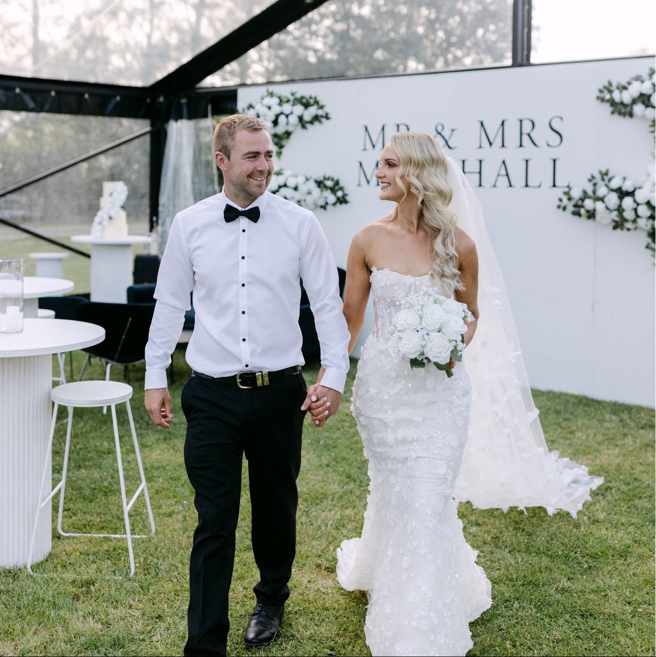 Wedding couple holding hands in front of a decorated backdrop with 'Mr & Mrs Marshall' text.