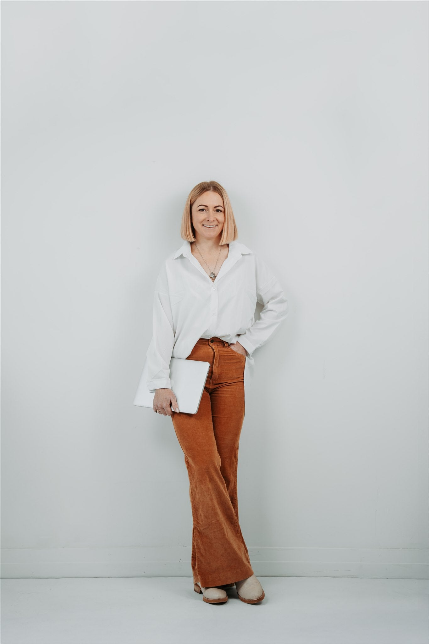 a styled shoot of a Woman wearing a white blouse and brown pants holding a Mac laptop against a light gray background